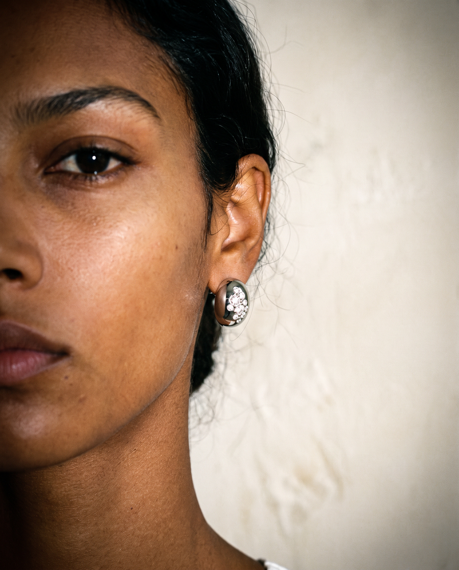 A close-up portrait of a woman wearing a small oval stud earring with a silver-toned metal border and a clear faceted center stone, set against a plain light beige wall.