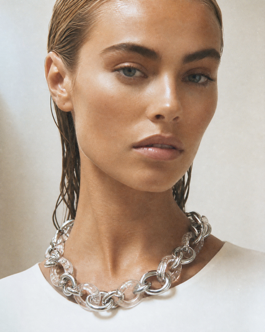 A close-up of a model wearing a chunky silver chain necklace with clear resin links, paired with a white top against a neutral beige background.