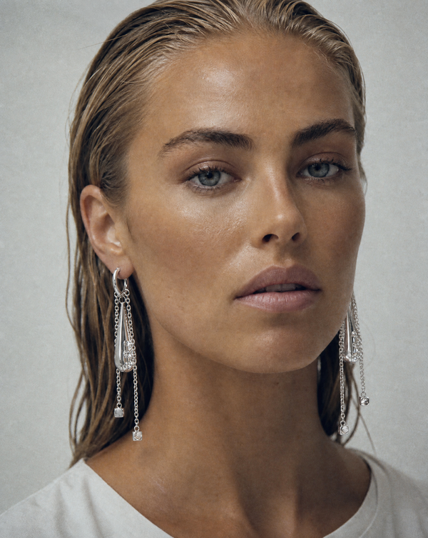 A close-up portrait of a woman with wet, slicked-back blonde hair wearing long, dangling silver and clear resin statement earrings with lab-grown diamonds, against a neutral light gray background.