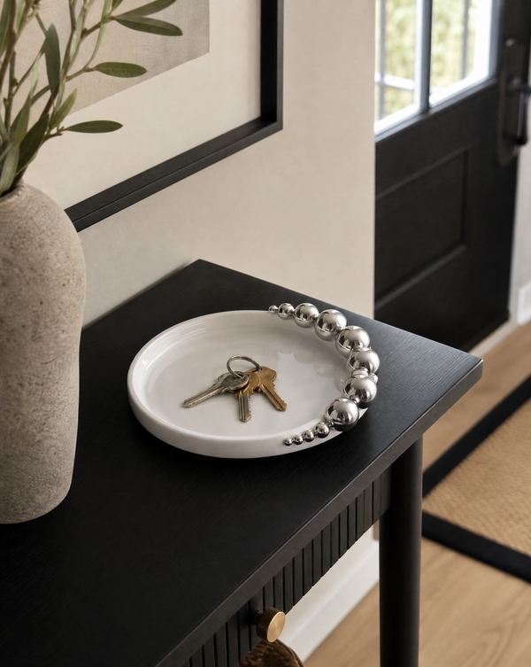 A white resin tray with a decorative silver beaded rim sits on a black console table in a modern entryway. 