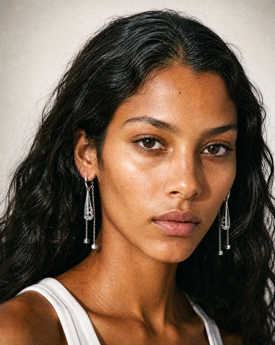 A close-up portrait of a woman with long dark hair wearing long silver dangle earrings with clear crystal-like drops against a soft beige background.