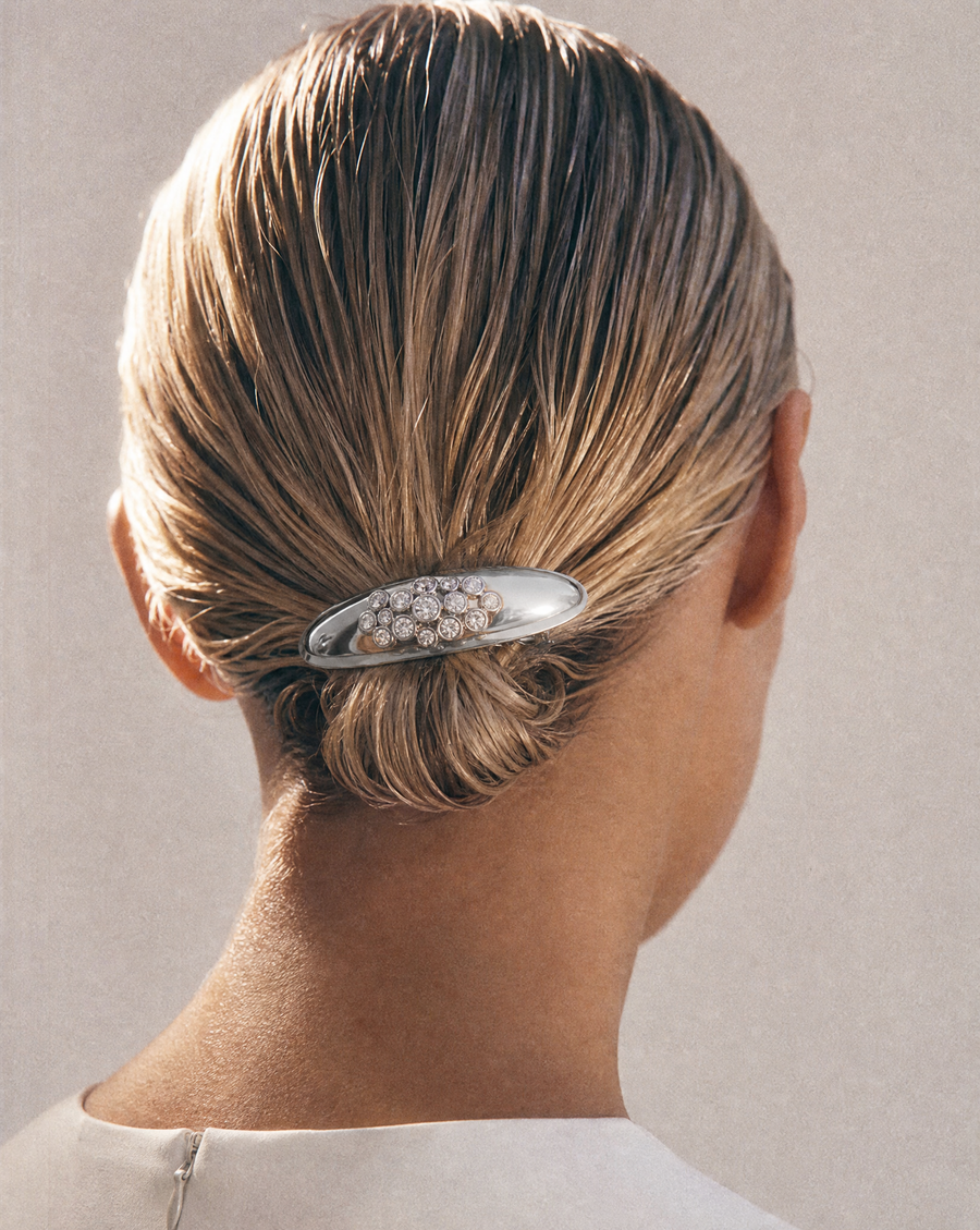 A close-up back view of a woman with her hair styled in a low bun, secured with a silver oval hair claw clip embellished with lab-grown diamonds against a neutral beige background.