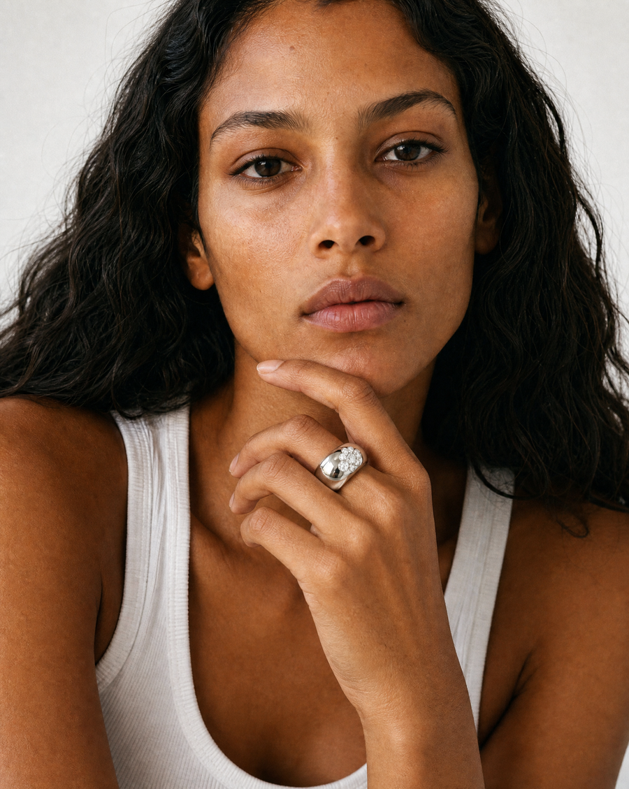 A close-up portrait of a woman wearing a large silver ring with round diamonds on her finger, paired with a white ribbed tank top against a plain light background.
