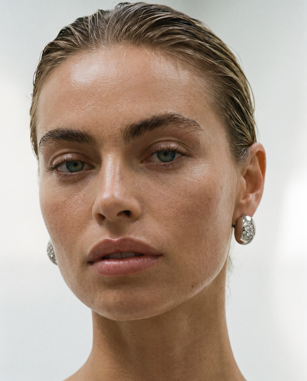 A close-up portrait of a woman with slicked-back hair wearing diamond bezel set silver hoop earrings against a plain light gray background.